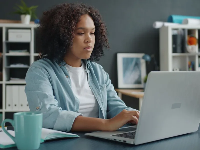 A young woman typing on a laptop at a desk in a home office with shelves and a notebook beside her